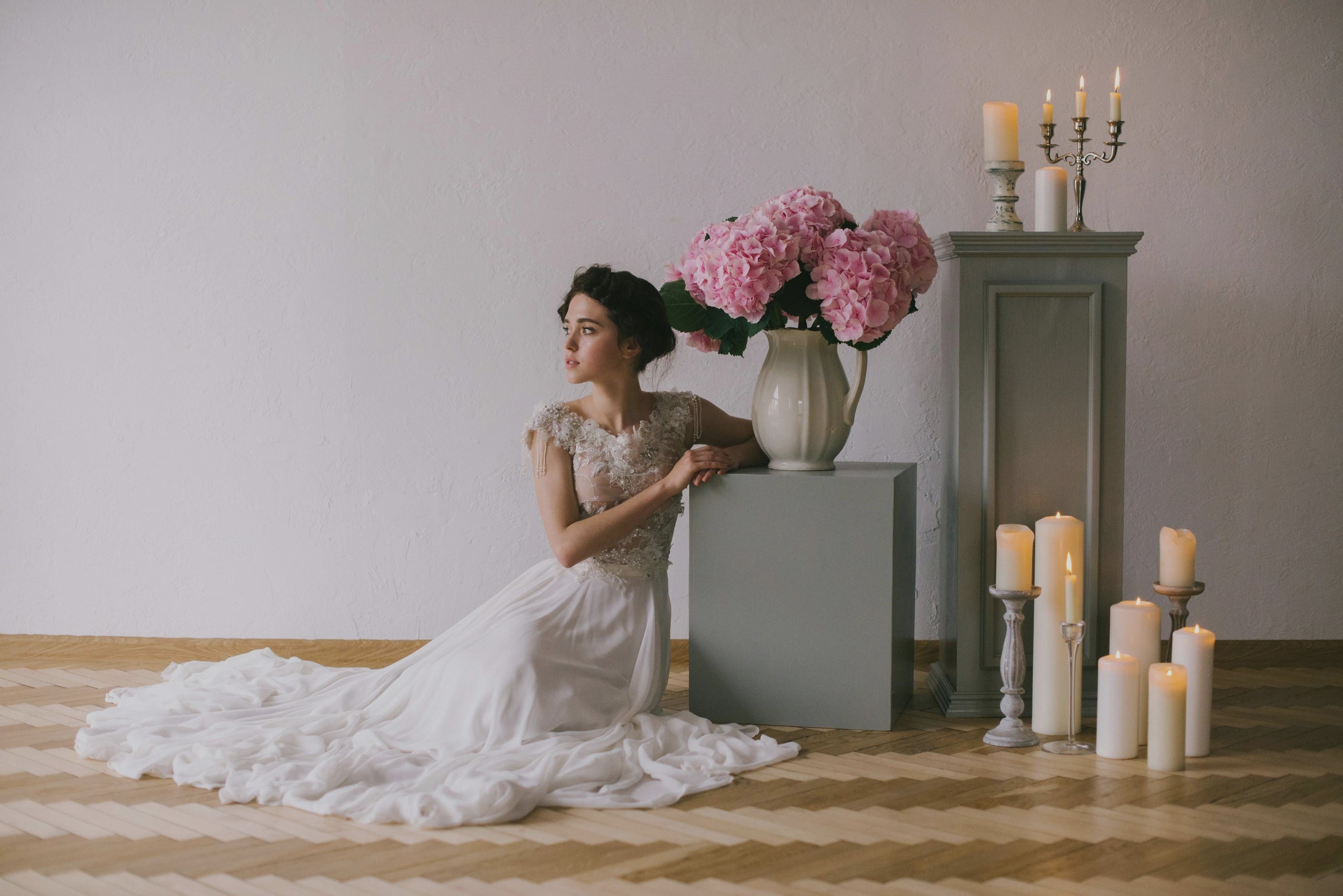 Young Caucasian women wearing a white wedding dress is sitting on the hardwood floor with her arm resting on a square platform near pink flowers in vase and several burning candles
