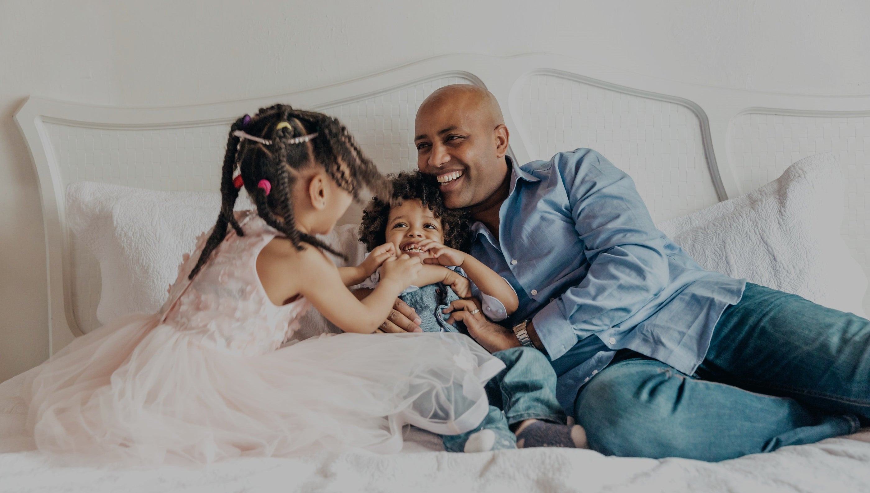 Middle-aged father and young daughter are tickling the toddler son on an all white bed in an all white room. All are laughing and smiling.