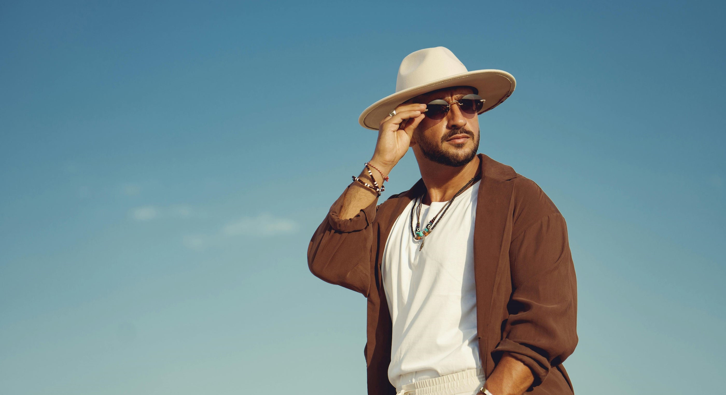 An international man wearing a wide brimmed sun hat, bracelets, necklaces, white shirt on the inside and brown shirt on the outside with blue skies in the background glances back as he holds onto his sunglasses.