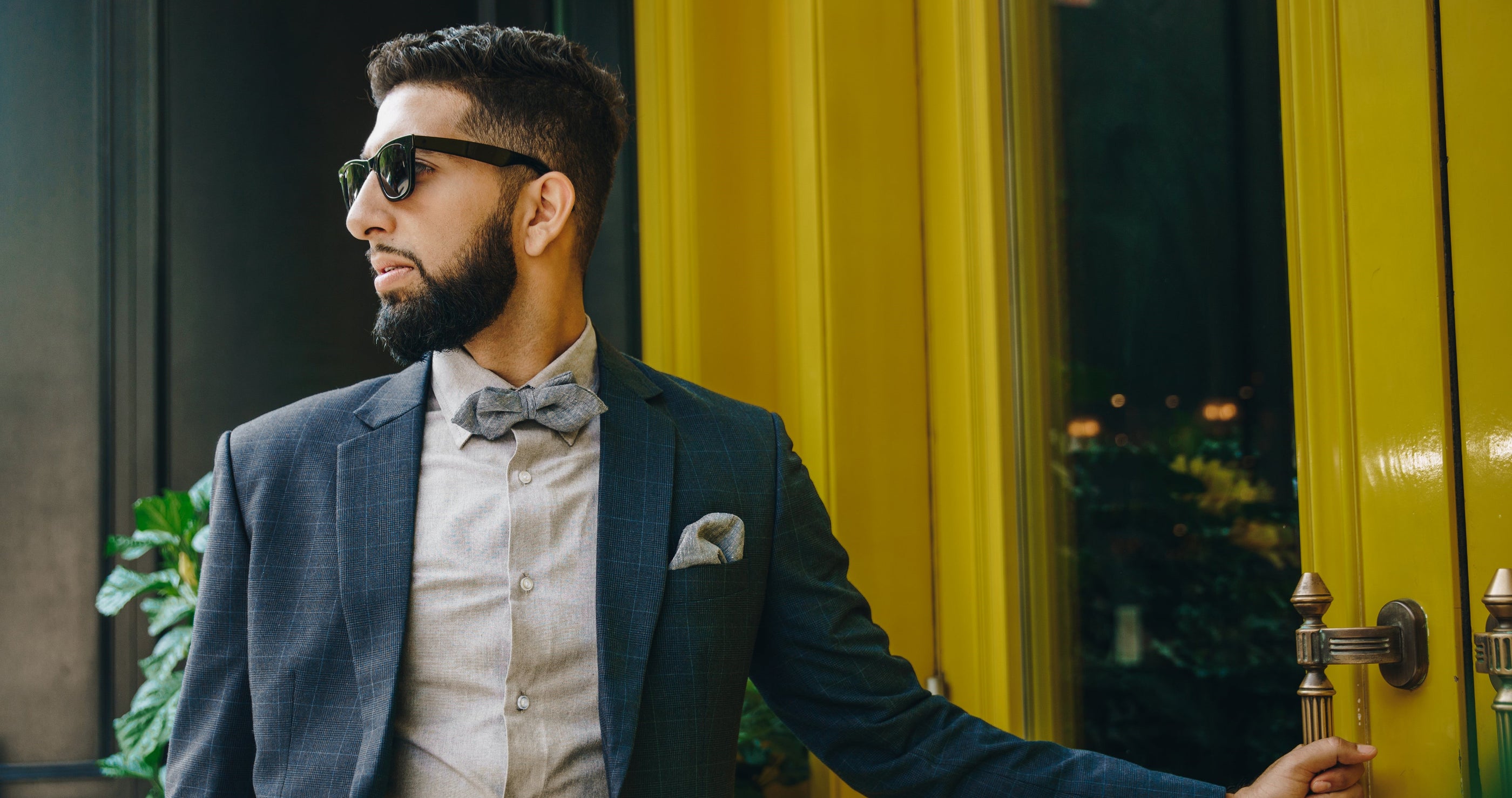 Dapper light skinned man with a suit, bow tie, and sunglasses is opening a grand yellow door.