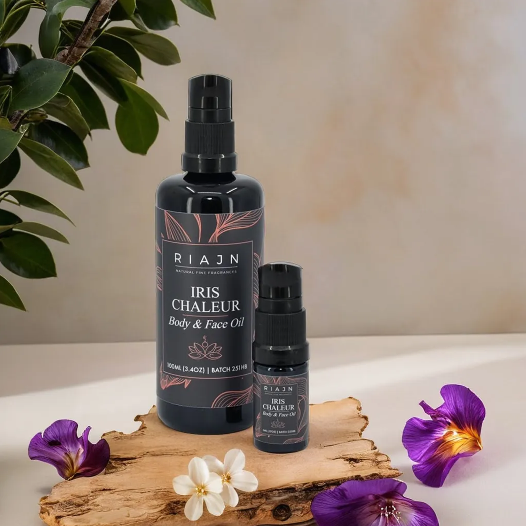 RIAJN Iris Chaleur Body & Face Oil bottles standing on a piece of driftwood next to purple iris flower pedals and small white pedals in a natural color room with a branch consisting of foliage showing in the upper left corner. Popular body oil and face oil for glowing skin.