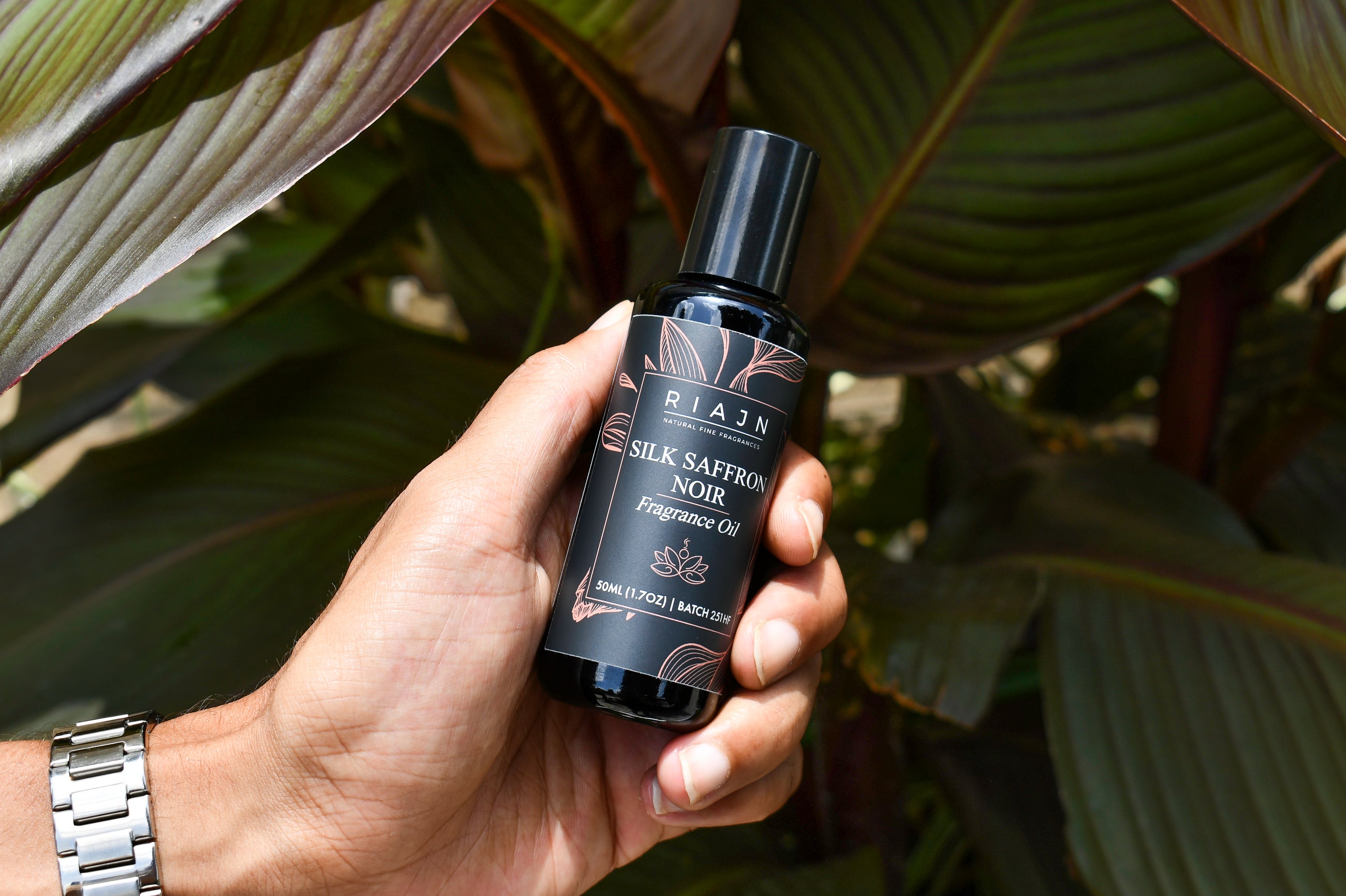 Hand holding a bottle of RIAJN Silk Saffron Noir perfume oil against a leafy background