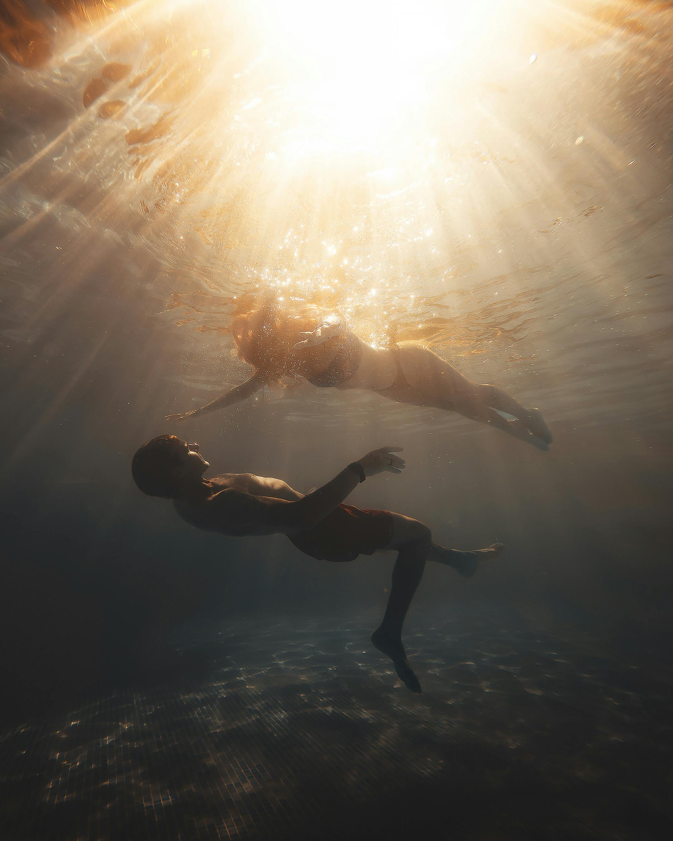 A couple floating towards each other with open arms in the deep ocean with bright sun rays shining on them through the top of the ocean.