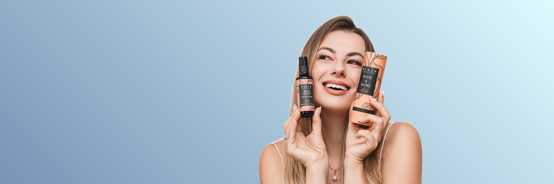 Women holding a riajn base scent bottle and tube packaging on the sides of her face and smiling. Hair & body mist, scent amplifier, layer base, long lasting EDP.