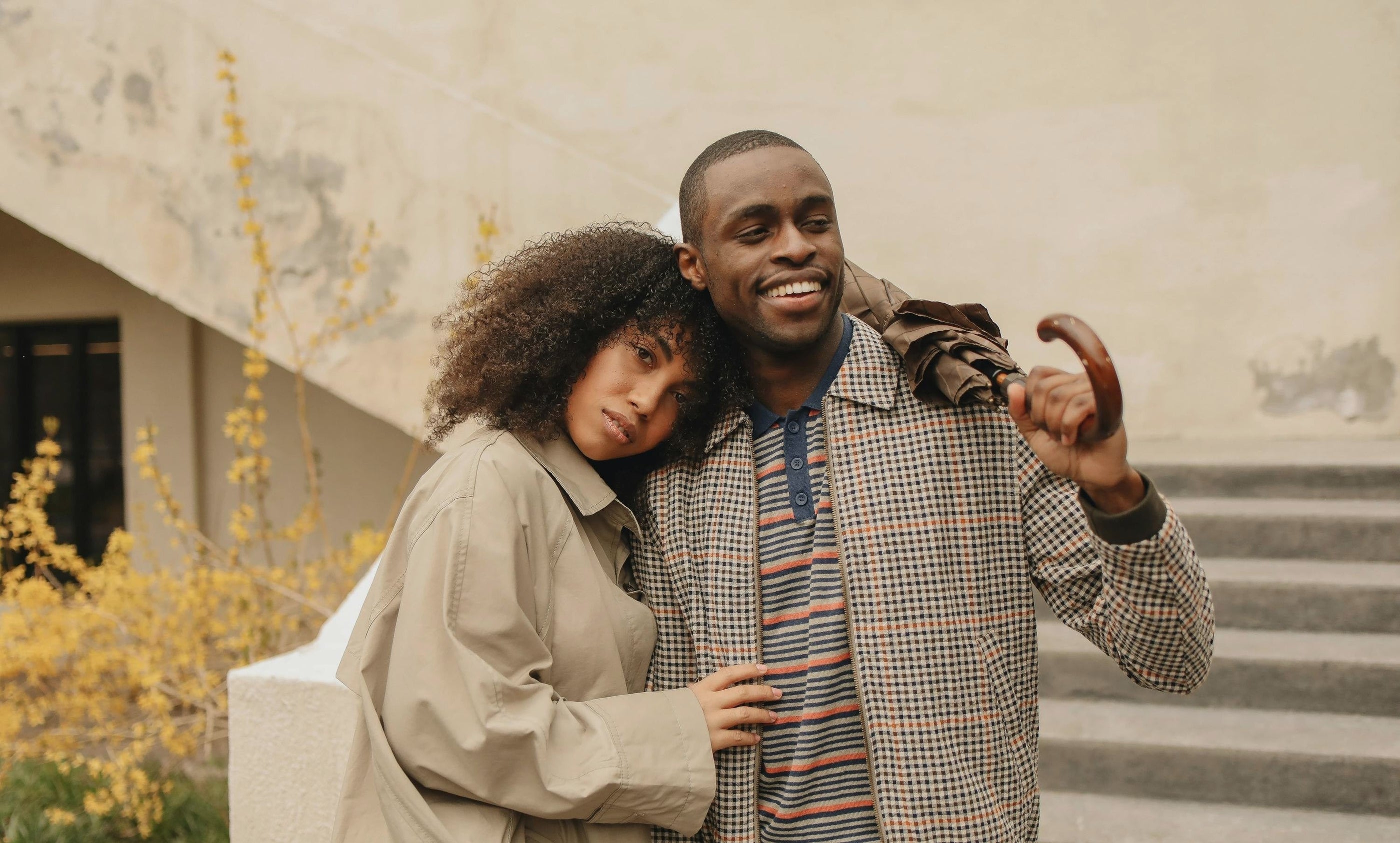 A couple wearing jackets with their cute pooch smiling drinking tea outdoors sitting on the ground with pillows leaning against a wooden fence.