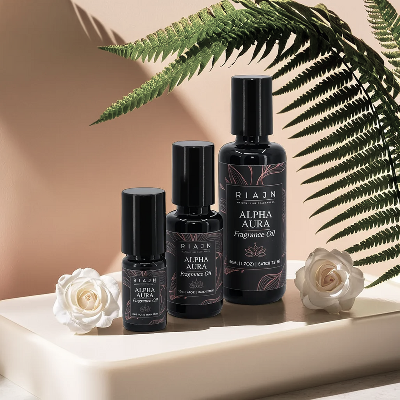 Set of RIAJAN Alpha Aura fragrance oil bottles with white roses and green fern leaves on a beige surface.