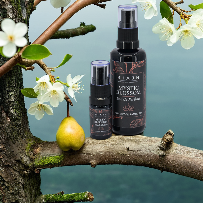 RIAJN Mystic Blossom Eau de Parfum bottles sitting on a tree branch near a hanging pear tree with blooming pear blossoms with mountain view in the background