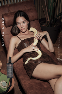 Woman sitting on a brown leather chair holding a snake, with 'Alpha Aura' branding.