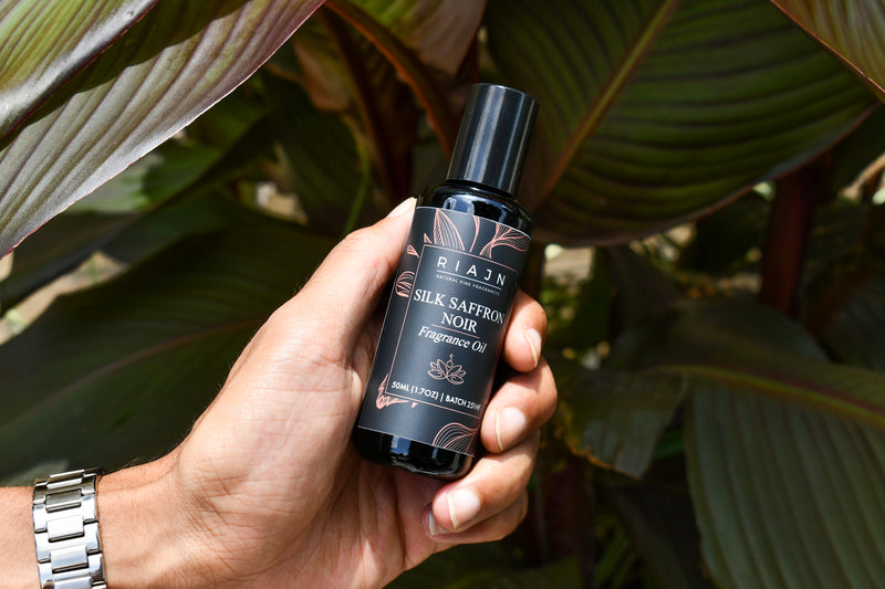 Hand holding a bottle of RIAJN Silk Saffron Noir perfume oil against a leafy background
