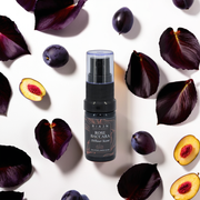 RIAJN Rose Baccara Diffuser Scent bottle laying on a white surface surrounded by dark purple rose pedals and plums with soft shadows.