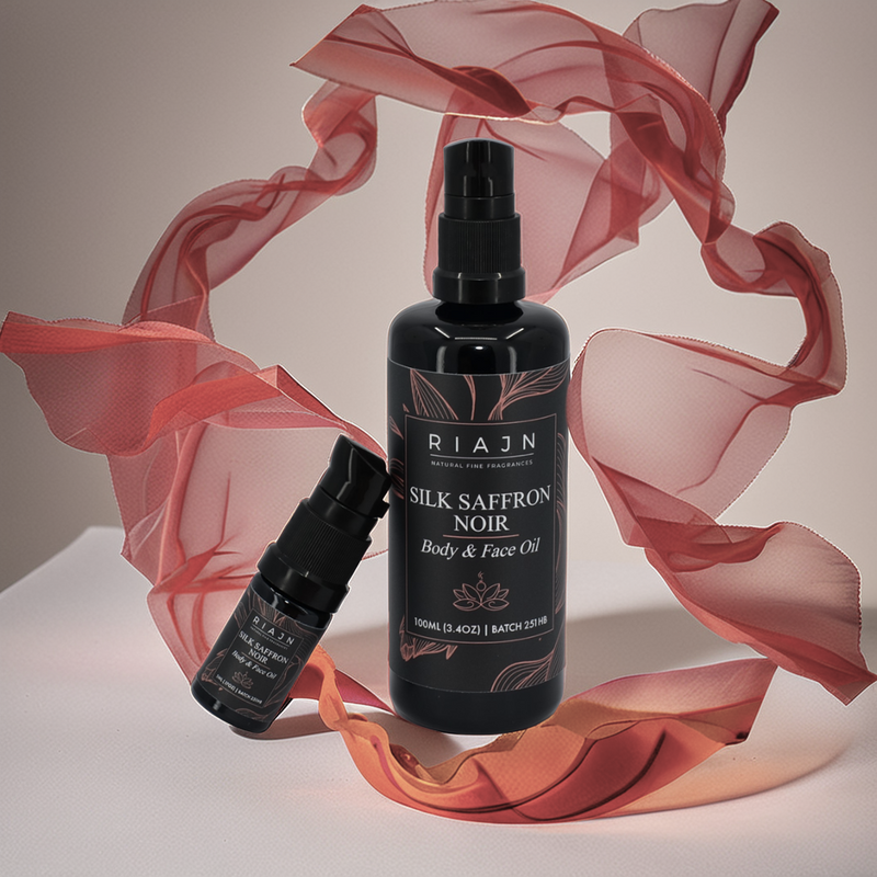 Two Riajn Silk Saffron Noir body and face oil bottles with red and orange silk fabric flowing in the background around the bottles.