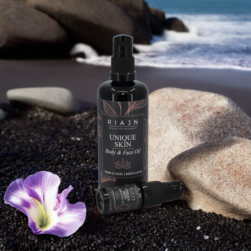 Riajn Unique Skin Body & Face Serum bottles on a beach with small black rocks and ocean in the background