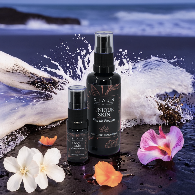 Two black RAIN Unique Skin perfume bottles with flowers on a wet surface with small wave of water splashing with ocean background